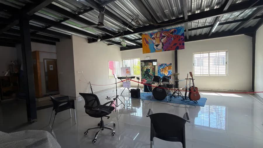 The full Hi-key Arts hall in daylight - polished white tile floor, black steel beams and a corrugated metal roof, with a keyboard, drum kit and guitars set up on a blue mat at the far wall beneath a large cheetah painting.