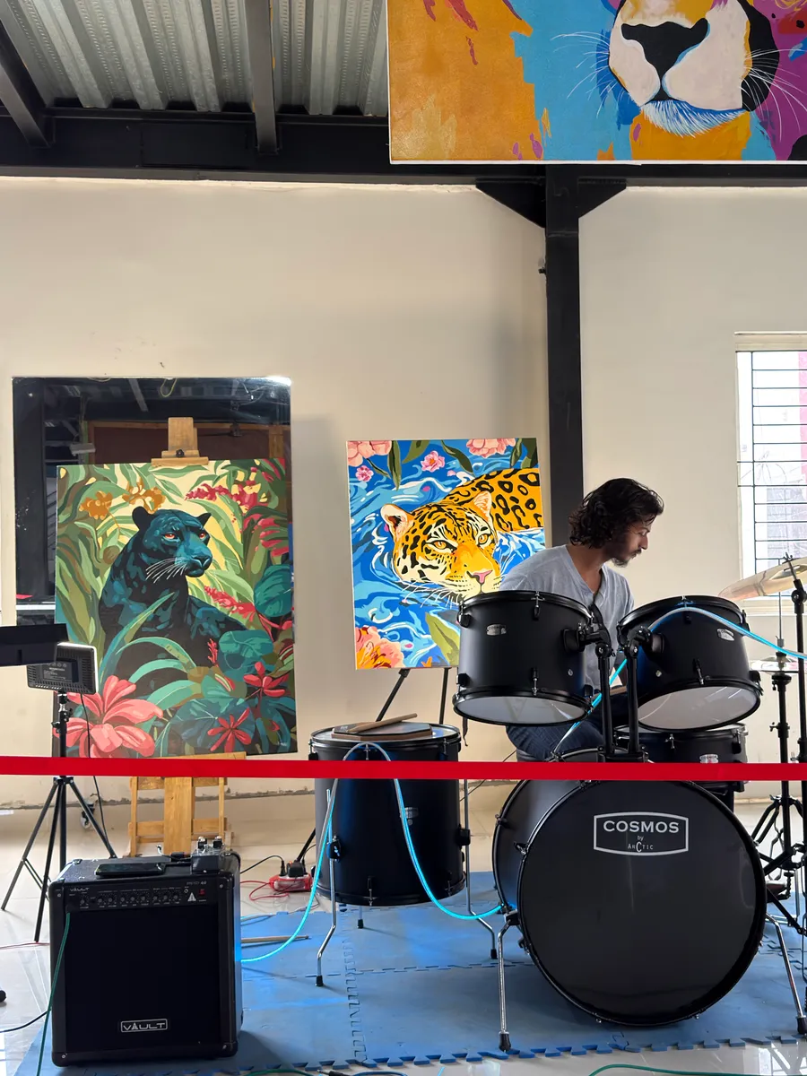 A drummer in a grey t-shirt playing the drum kit in daylight, with a black panther painted among jungle leaves standing on a wooden easel and a jaguar drifting in blue water on the canvas beside him.